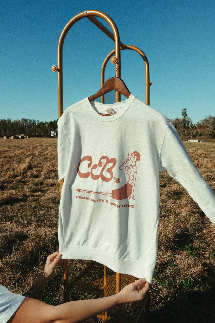 CB's white and dusty rose logo crewneck on a hanger in a field